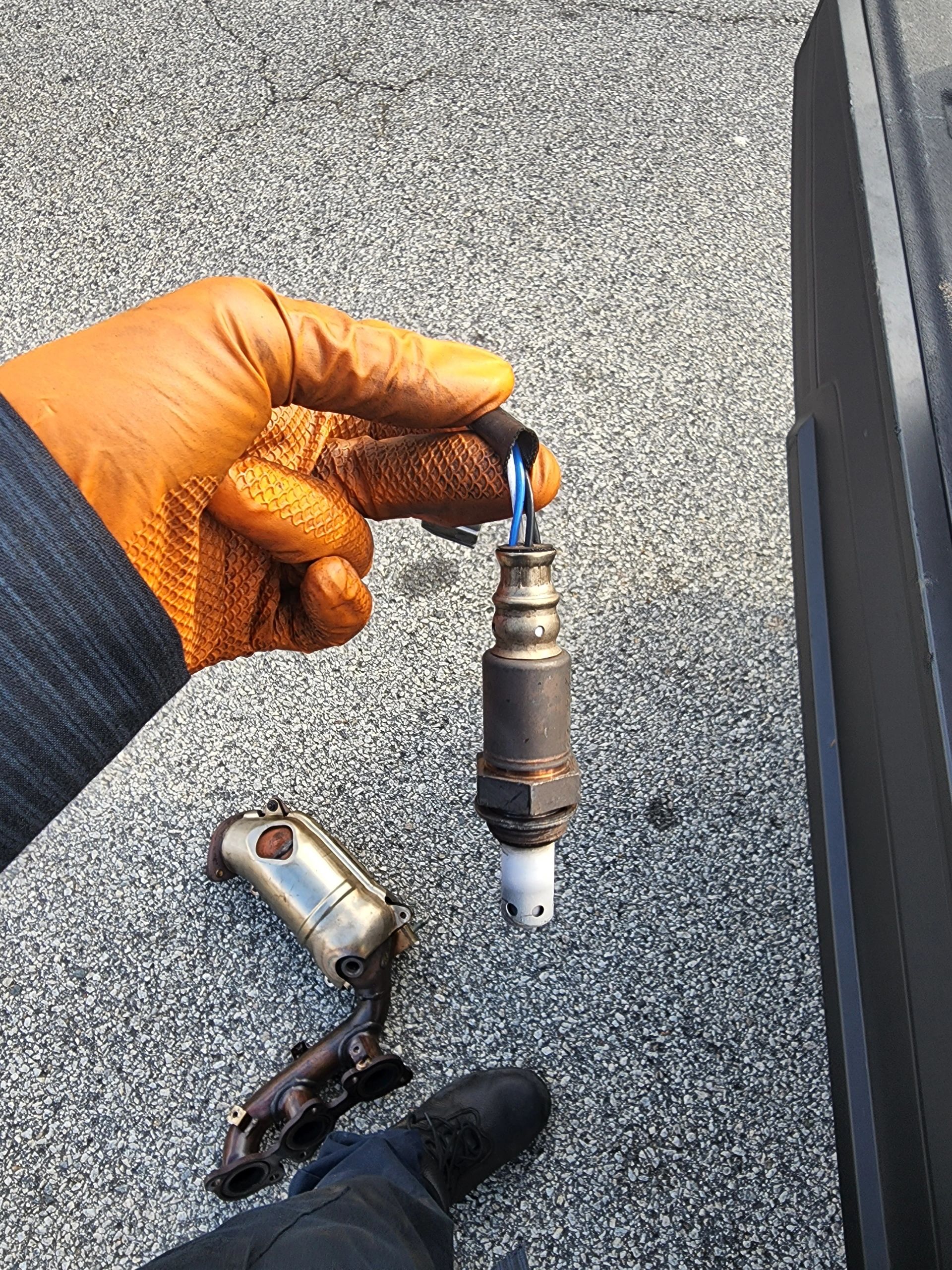 Tech On The Way — mechanic in orange glove holding a failed oxygen sensor next to the catalytic converter elbow during an on-site replacement