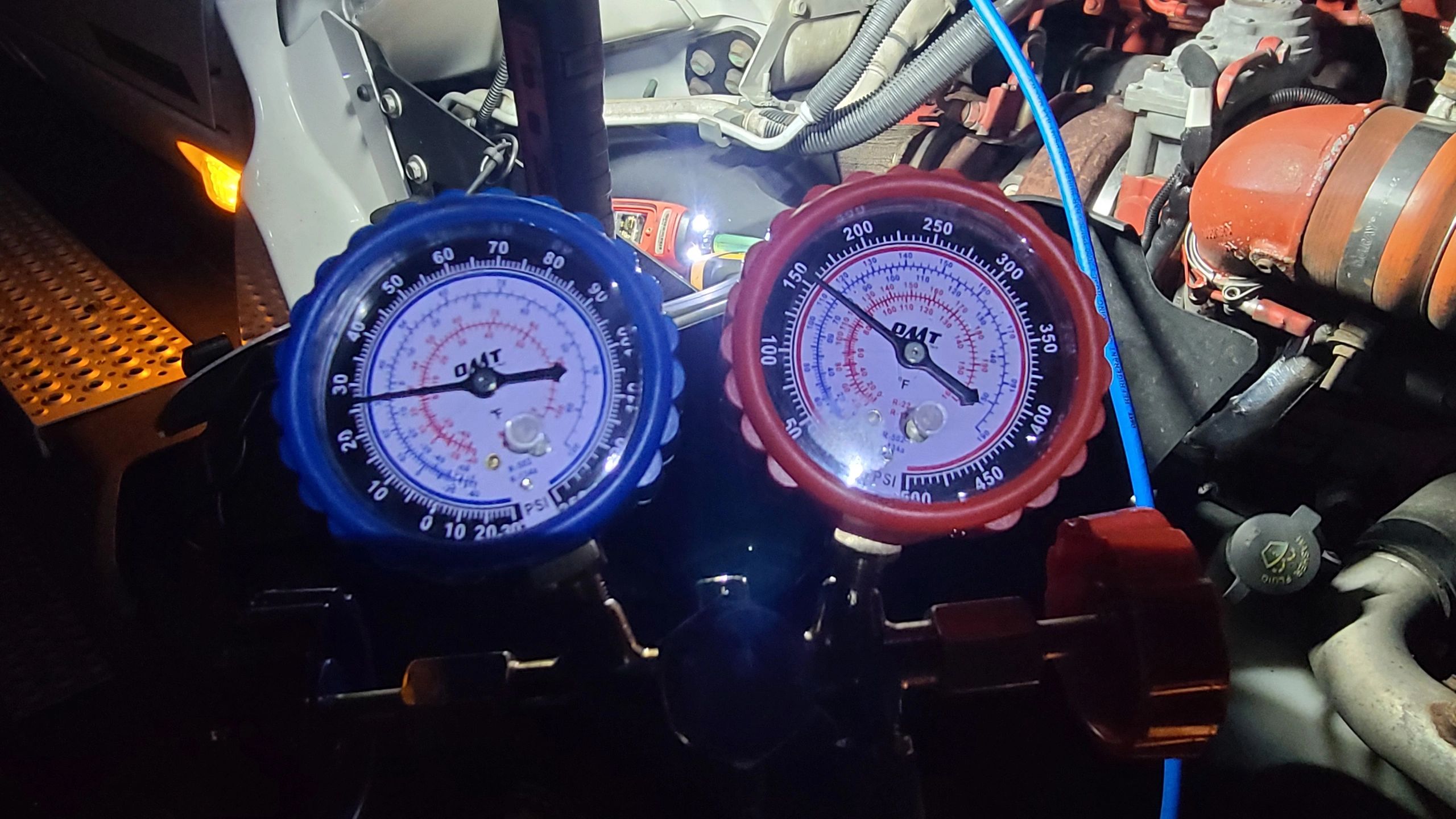 Tech On The Way — blue and red refrigerant manifold gauges mid-diagnostic showing low-side and high-side pressure readings in an engine bay